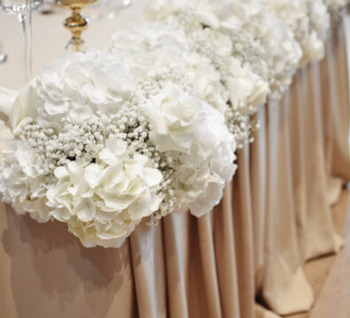 dekoracja beżowa na wesele, stolik z beżową narzutą i białymi dużymi hortensjami.