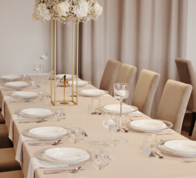 dekoracja stolika dla gości beżowym obrusem, białymi serwetami, wysokim stojakiem z kwiatami w białym kolorze. hortensje, róże, gibsówka